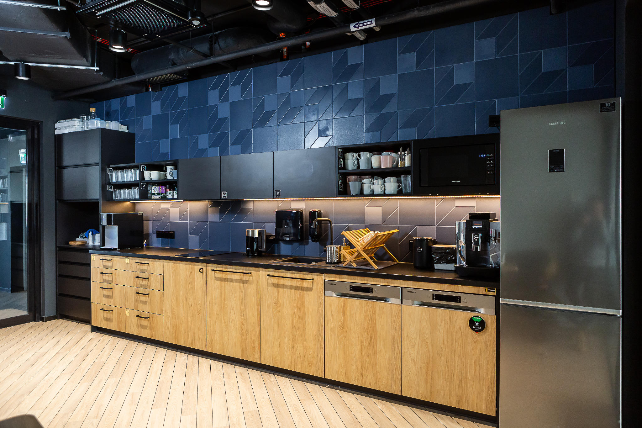 Nowoczesna kuchnia biurowa z drewnianymi frontami szafek, czarnym blatem i niebieską mozaiką na ścianie, wyposażona w srebrną lodówkę Samsung i ekspresy do kawy.