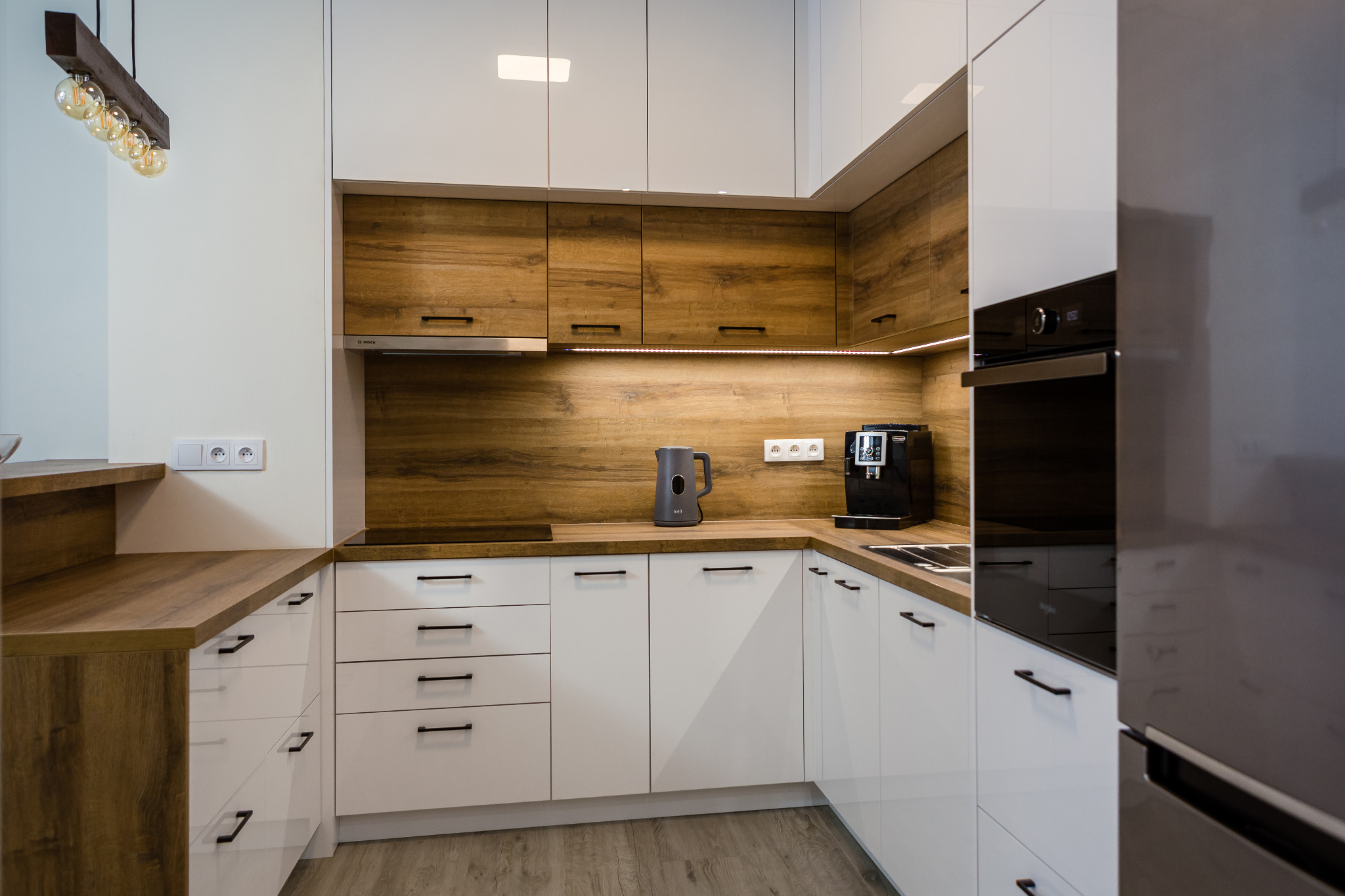 Nowoczesna kuchnia z białymi lakierowanymi frontami dolnych szafek i drewnopodobnymi górnymi szafkami, oświetleniem podszafkowym LED, czarnym piekarnikiem i ekspresem do kawy na blacie.