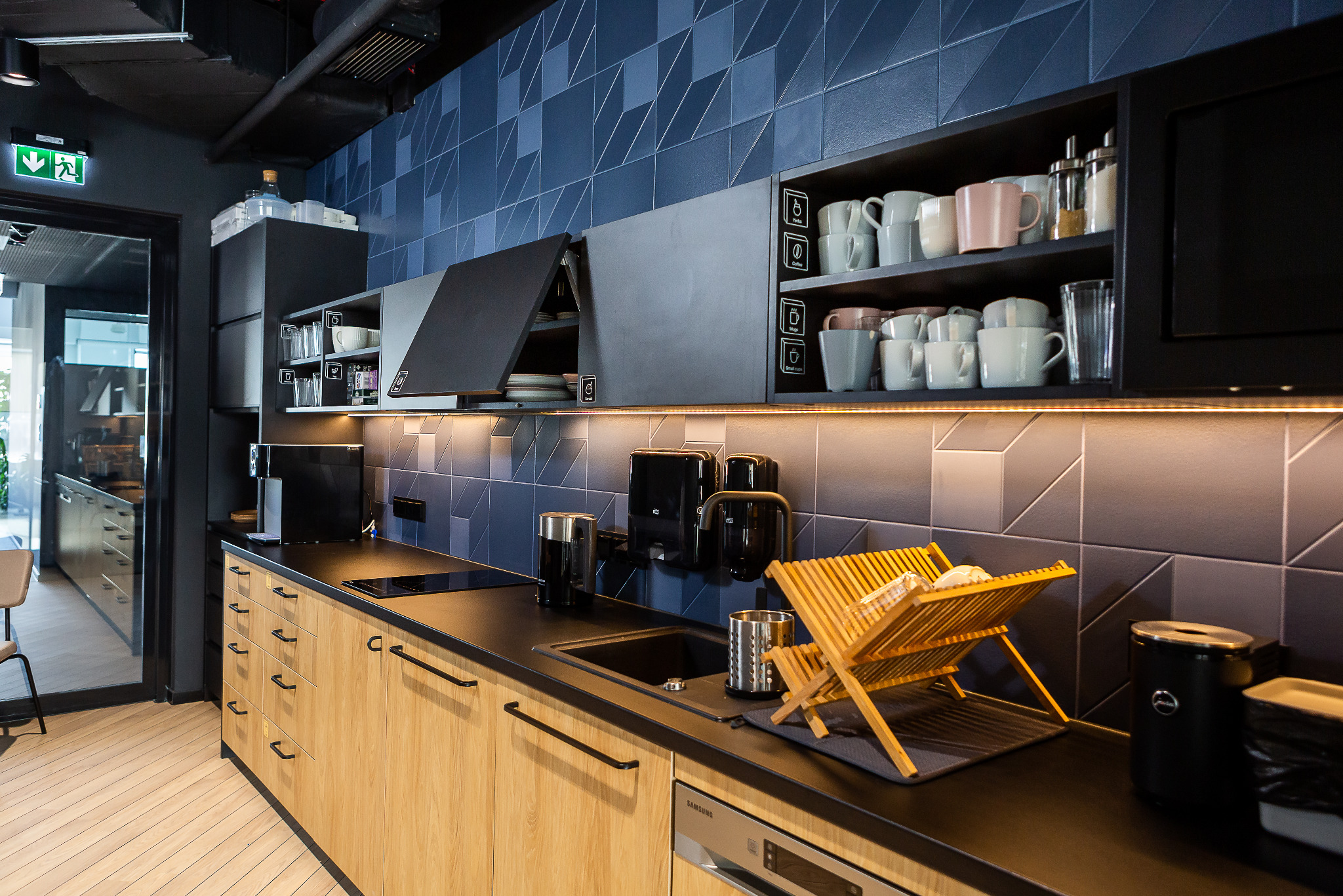 Nowoczesna kuchnia z drewnianymi frontami szafek, czarnym blatem i geometrycznymi płytkami na ścianie, wyposażona w zlew, suszarkę na naczynia, ekspres do kawy i podświetlane szafki górne...