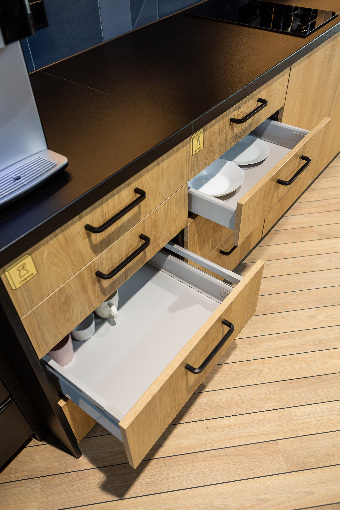 Nowoczesna zabudowa kuchenna z drewnianymi frontami, czarnym blatem i otwartymi szufladami, w których widoczne są naczynia i akcesoria kuchenne. Na frontach szuflad umieszczone są piktogramy.