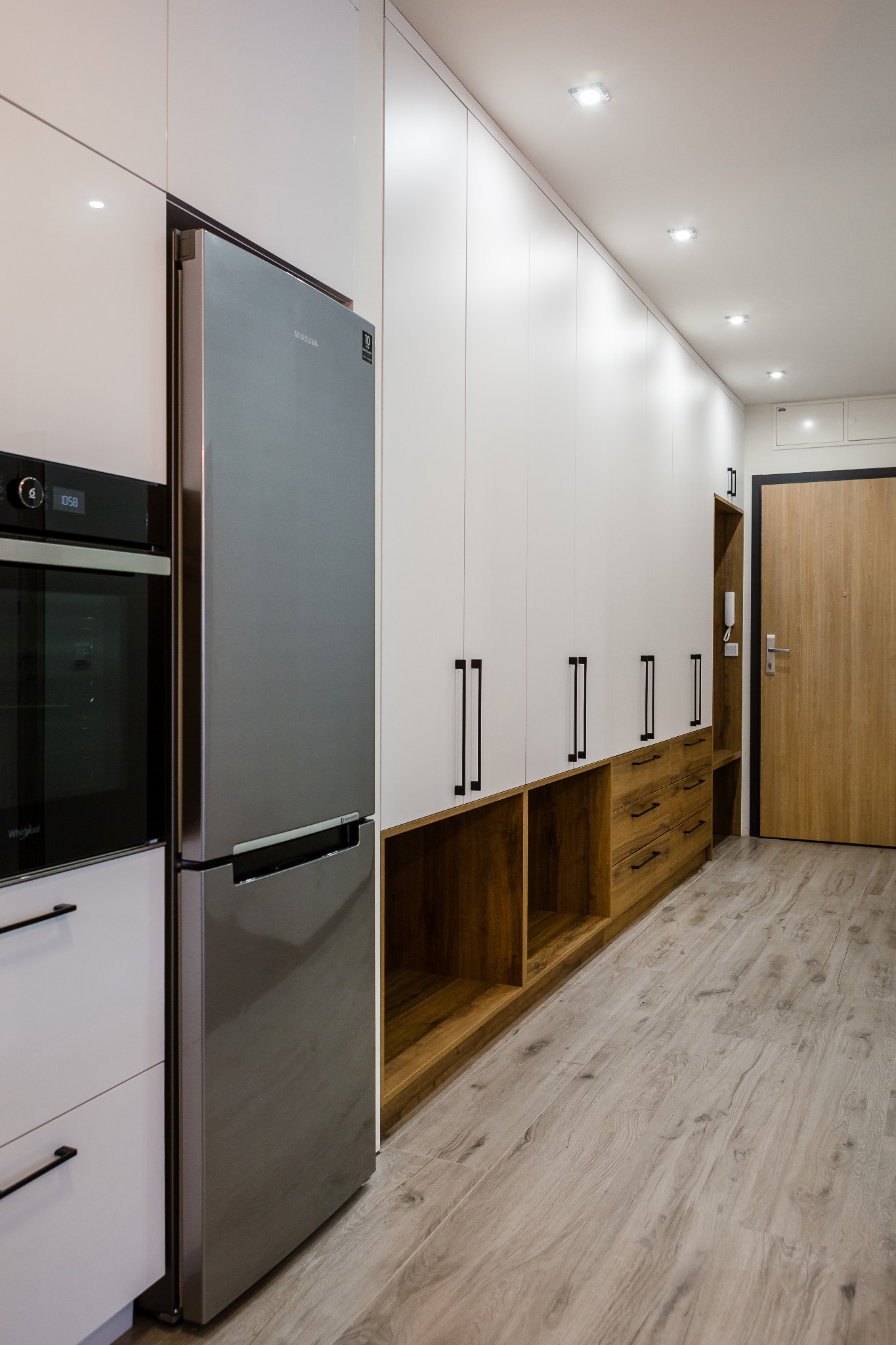 Długa zabudowa meblowa z białymi szafkami, drewnianymi szufladami i wnękami, zintegrowana z lodówką i piekarnikiem w nowoczesnym mieszkaniu, oświetlenie punktowe w suficie.