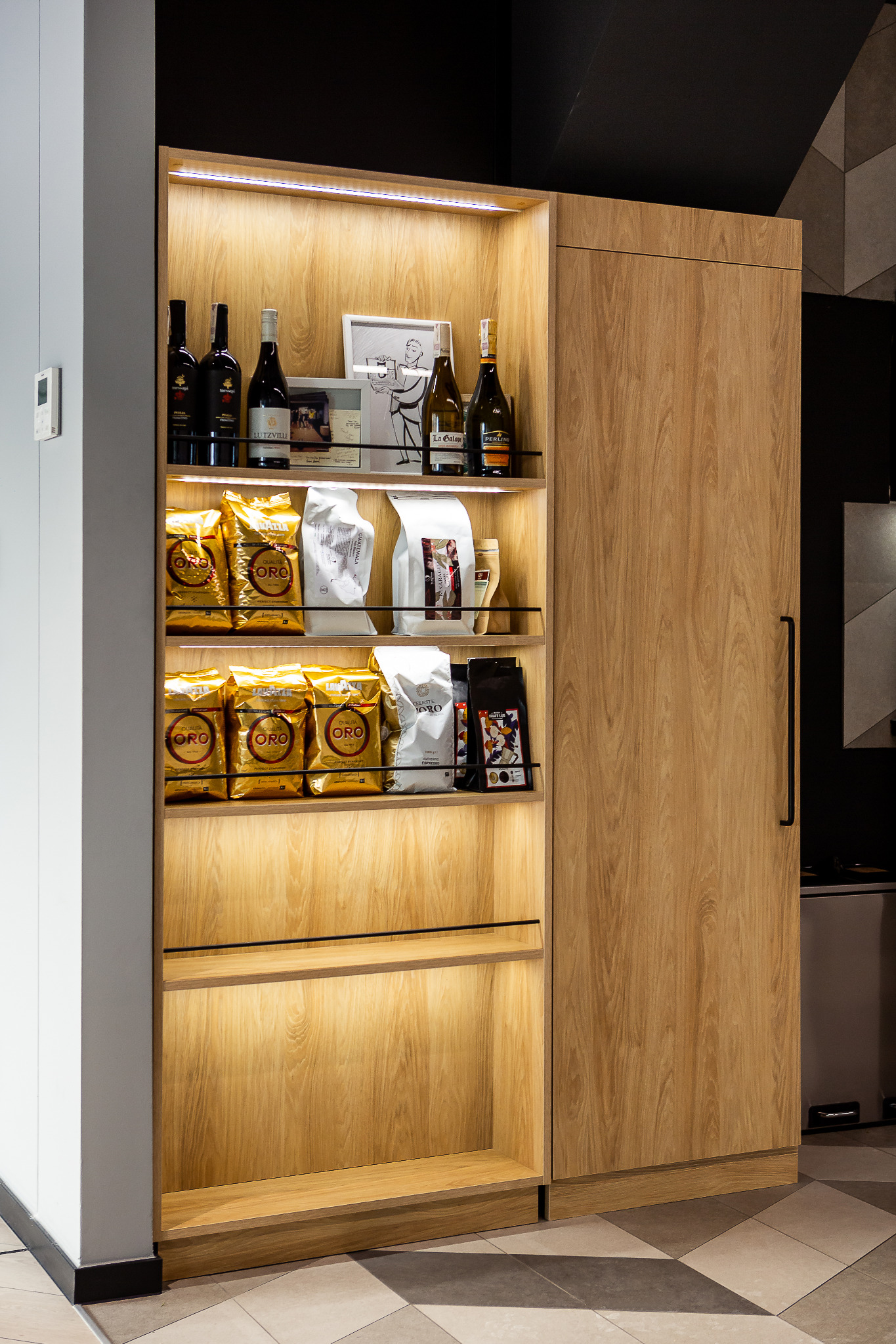 Wysoka, drewniana szafa wnękowa z oświetleniem LED, wypełniona zapasami kawy Qualita Oro, win i dekoracyjnych przedmiotów, na tle podłogi w geometryczny wzór.