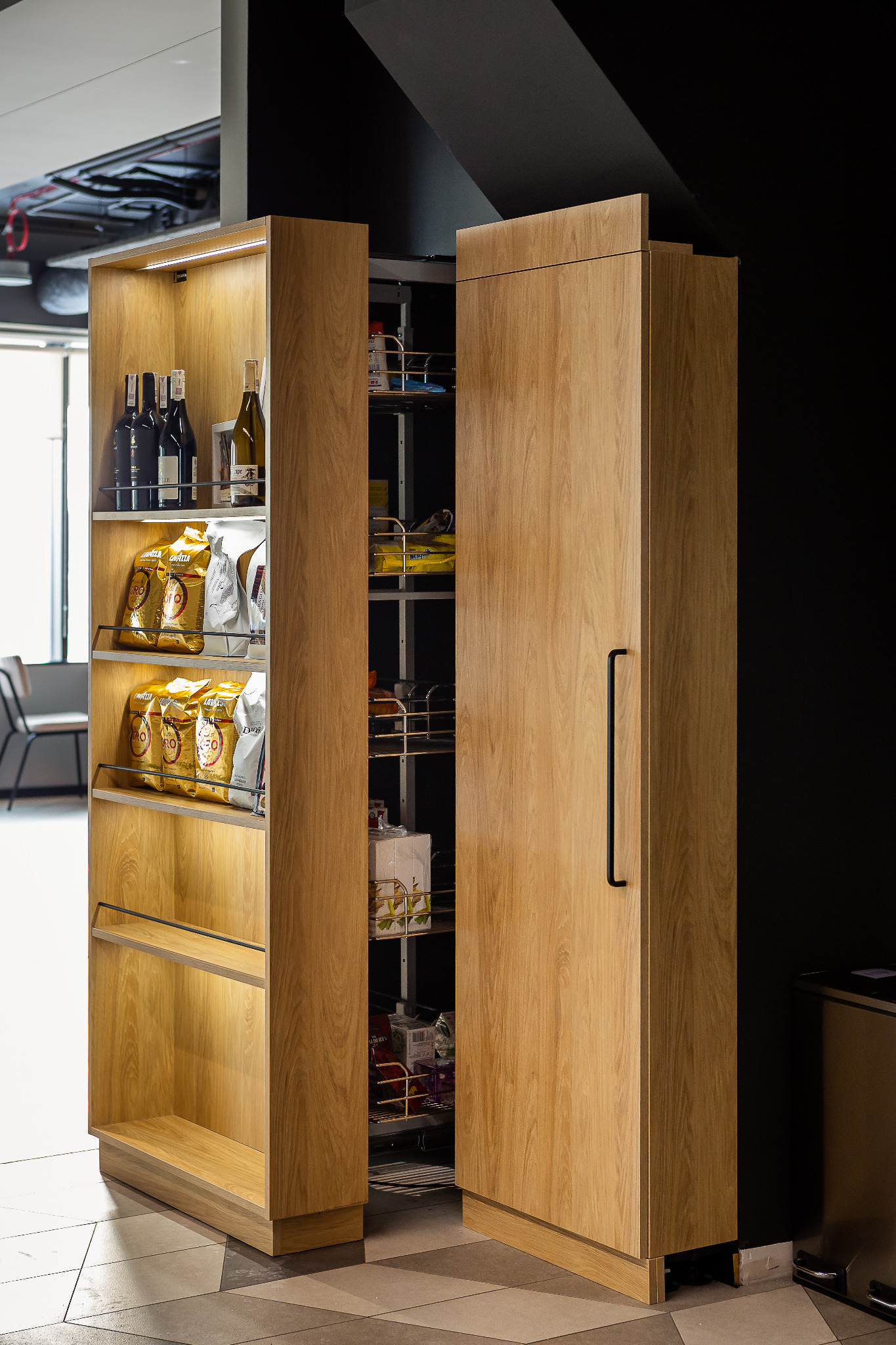 Otwarta szafka cargo z drewnianymi frontami i metalowymi półkami, wypełniona produktami spożywczymi, obok regał z winami i kawą, oświetlenie LED wewnątrz.