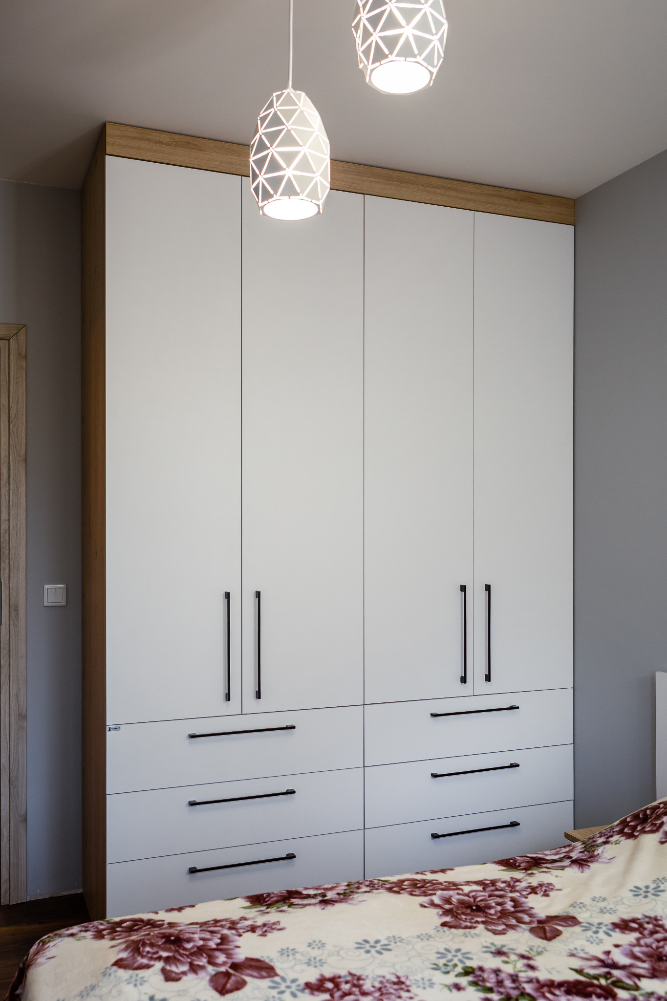 Biała szafa wnękowa z drewnianym akcentem, czarnymi uchwytami i dwiema lampami wiszącymi w kształcie geometrycznym, widoczny fragment łóżka z kwiecistą pościelą.