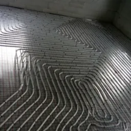 Białe rury ogrzewania podłogowego ułożone spiralnie na izolacji z siatką w surowym wnętrzu z oknem, widok z góry pod kątem.