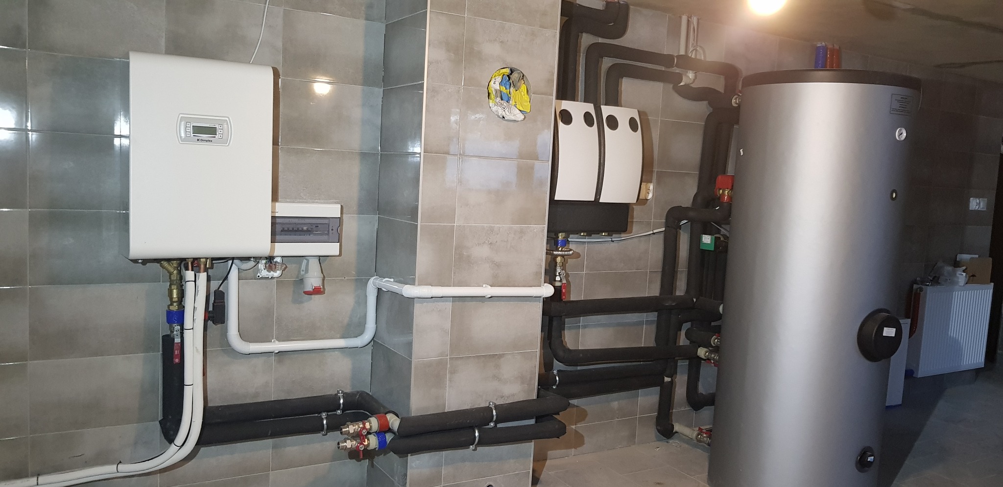 Instalacja pompy ciepła z zasobnikiem ciepłej wody użytkowej i rozbudowanym systemem rur w pomieszczeniu wykończonym szarymi płytkami.