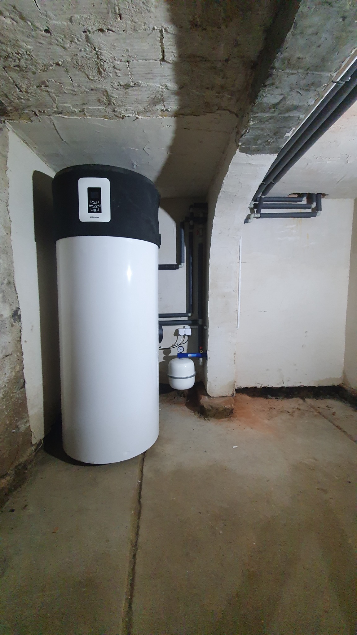 Biały zbiornik akumulacyjny z czarną górną częścią i wyświetlaczem, zainstalowany w piwnicy z betonowym sufitem i rurami instalacyjnymi.
