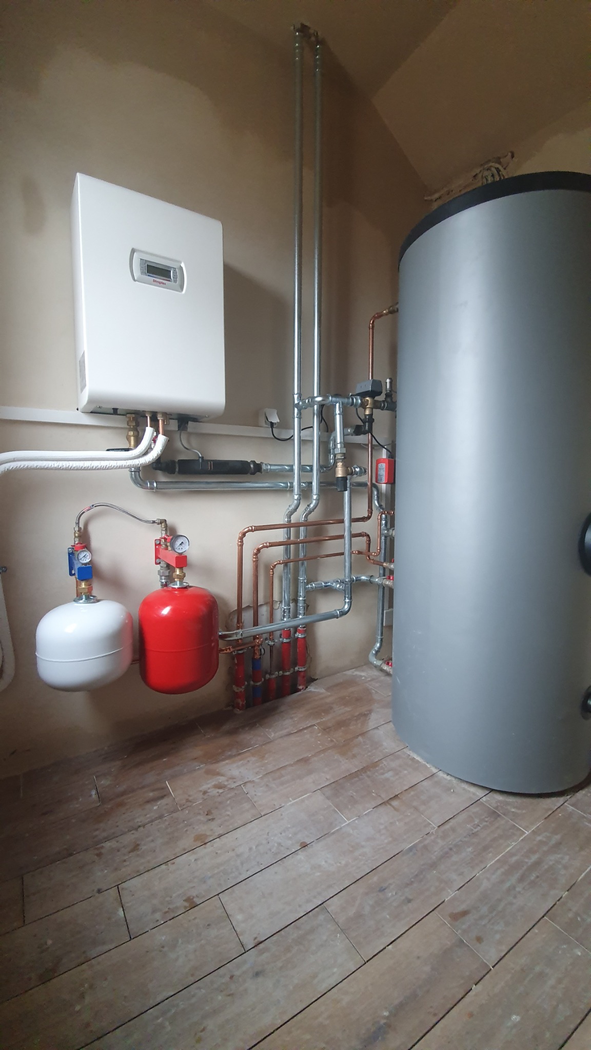 Instalacja grzewcza z białym kotłem, szarym zasobnikiem ciepłej wody oraz czerwonym i białym naczyniem przeponowym, widoczne liczne rury miedziane i stalowe, zamontowana w pomieszczeniu z podłogą...