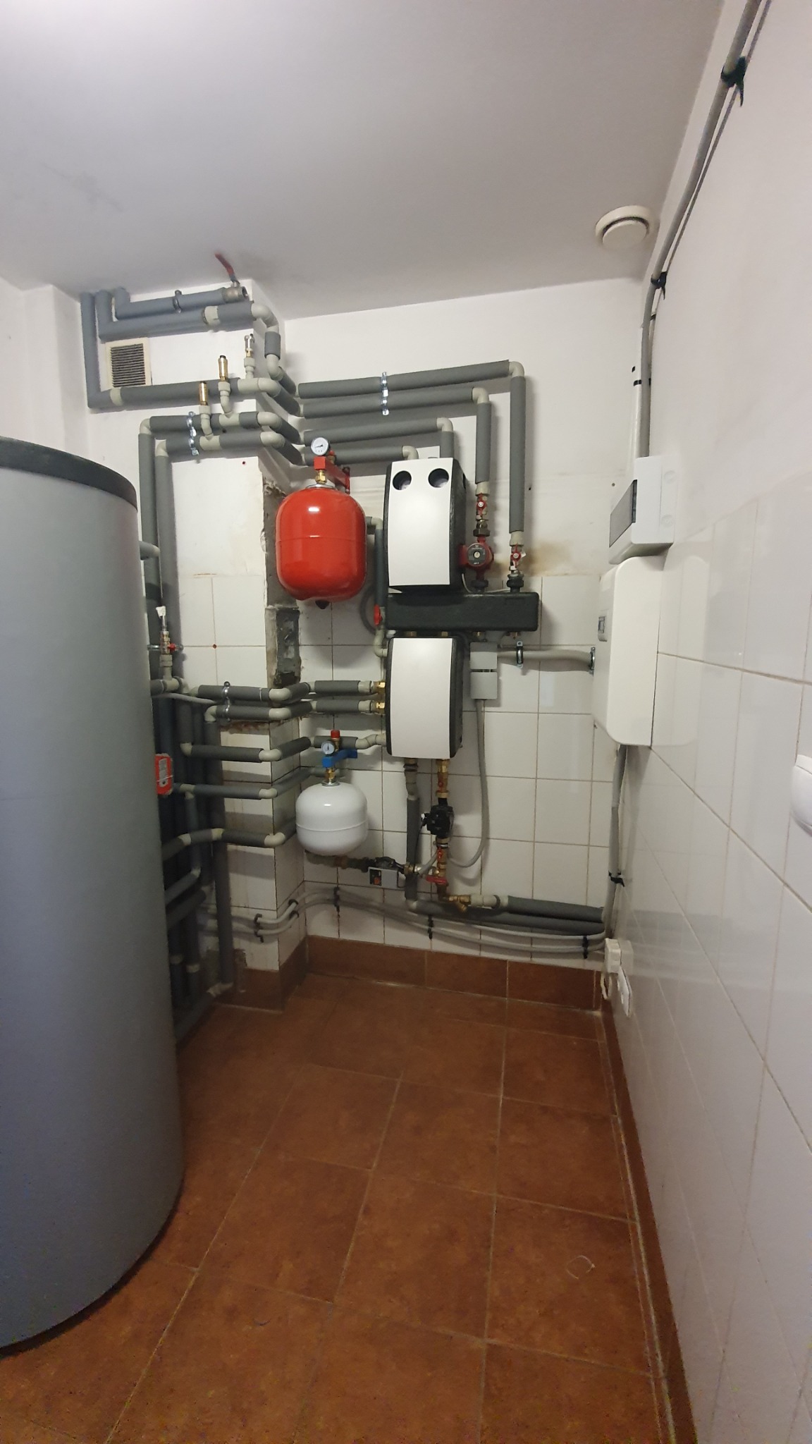 Węzeł grzewczy z szarymi rurami izolowanymi termicznie, czerwonym zbiornikiem wyrównawczym, białym zbiornikiem i dwoma białymi urządzeniami na ścianie wyłożonej białymi płytkami.