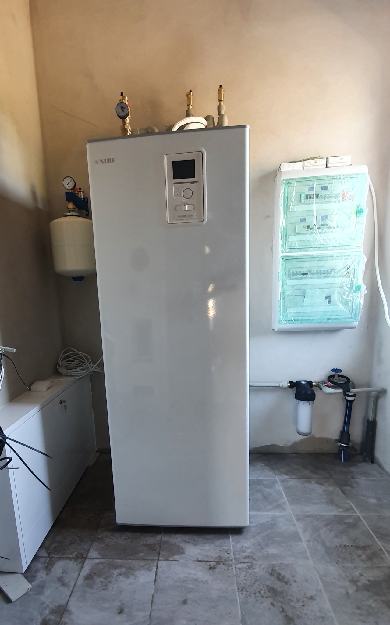 Biała pompa ciepła Nibe z podłączonymi rurami, manometrami i zbiornikiem wyrównawczym, obok skrzynka bezpiecznikowa w folii ochronnej i filtr wody na tle beżowej ściany i szarej podłogi.