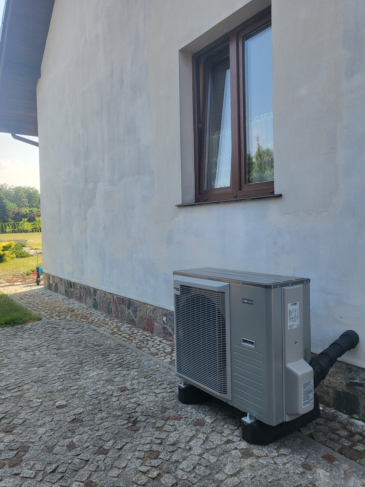 Szara jednostka zewnętrzna urządzenia grzewczego na czarnych podstawkach, zamontowana przy ścianie domu z kamienną podmurówką i brukowanym chodnikiem, widoczna instalacja odprowadzająca.