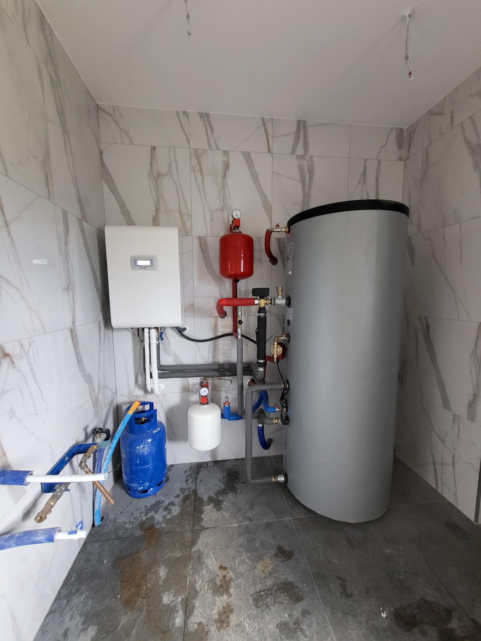 Instalacja grzewcza z zasobnikiem i białą jednostką sterującą na marmurowych płytkach, widoczna butla gazowa i elementy instalacji hydraulicznej.