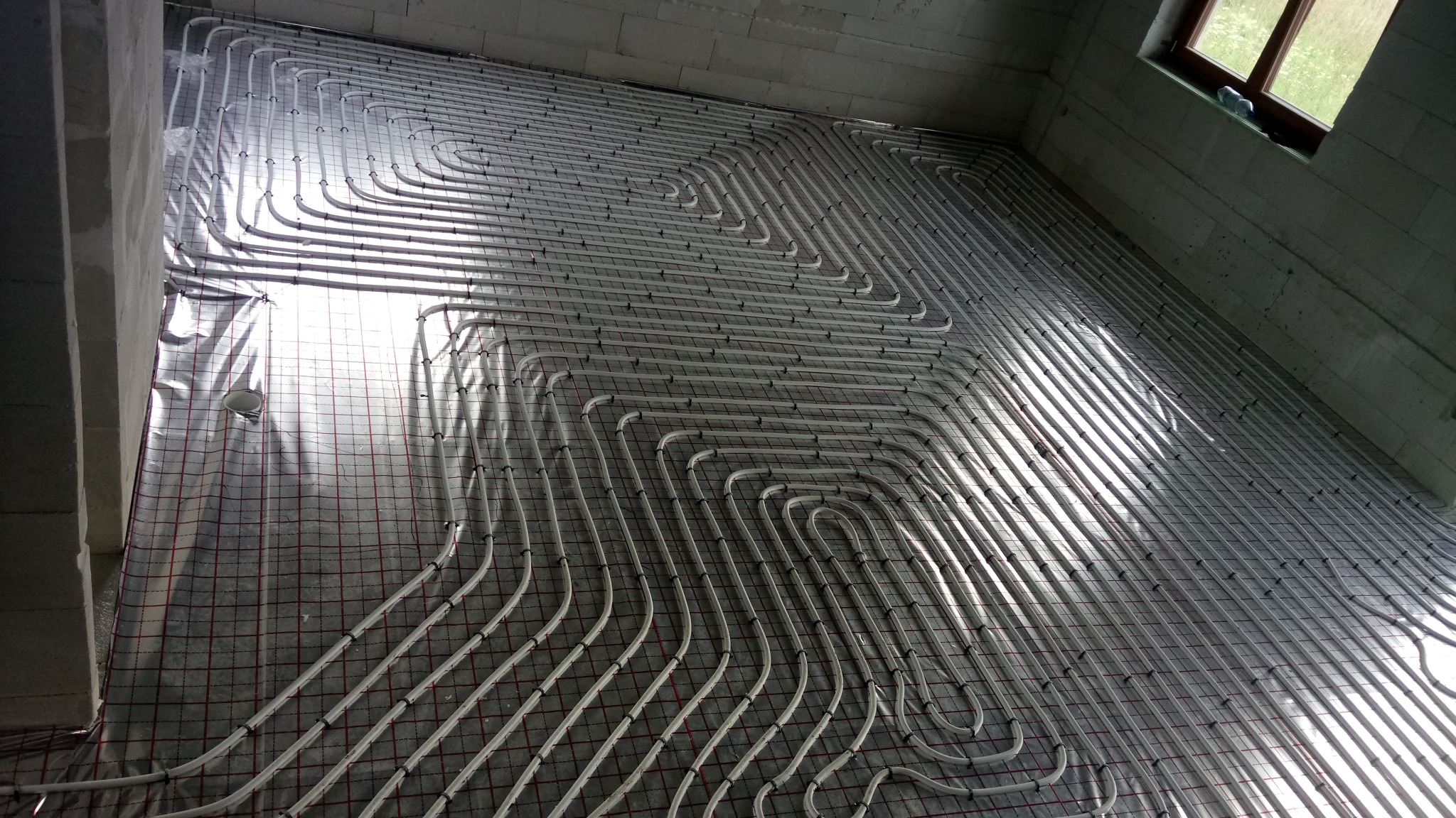Białe rury ogrzewania podłogowego ułożone spiralnie na izolacji z siatką w surowym wnętrzu z oknem, widok z góry pod kątem.