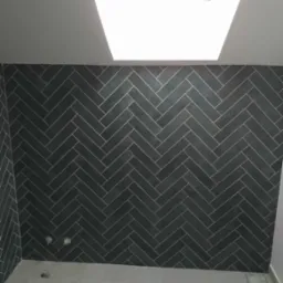 Ciemnoszara ściana wyłożona płytkami w jodełkę z jasnymi fugami, oświetlona przez okno dachowe, w nowej łazience z jasną podłogą.