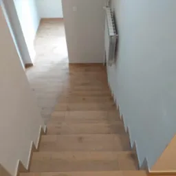 Schody wyłożone panelami imitującymi drewno, z widocznym grzejnikiem na ścianie i fragmentem korytarza w tle.