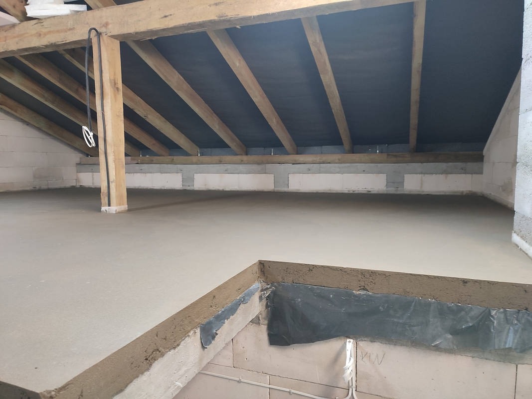 Świeżo wylana, gładka posadzka w surowym stanie poddasza z widoczną konstrukcją drewnianą dachu i ścianami z bloczków betonowych.