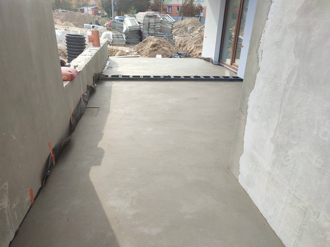 Świeżo wylany, gładki jastrych na tarasie domu jednorodzinnego, widoczne przygotowania do dalszych prac wykończeniowych, w tle stosy materiałów budowlanych.