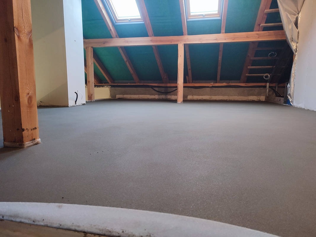 Świeżo wylana, gładka wylewka w poddaszowym pomieszczeniu z drewnianą konstrukcją dachu i oknami połaciowymi.