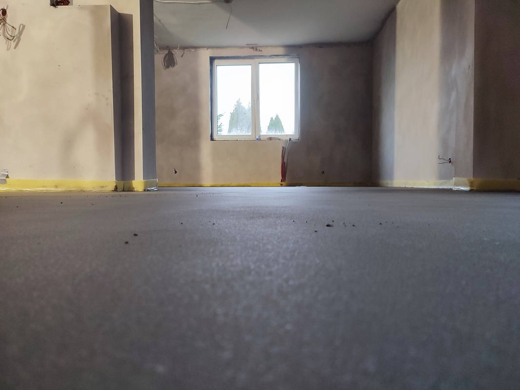 Wylany, świeży jastrych w pomieszczeniu z oknem, zabezpieczony żółtą taśmą przy ścianach.