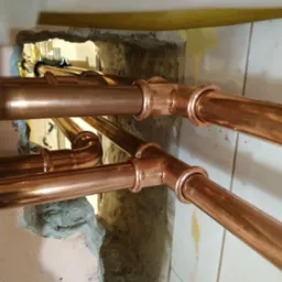 Miedziane rury instalacyjne, prowadzone przez otwór w ścianie, widoczne połączenia lutowane, surowy stan wykończenia ściany.