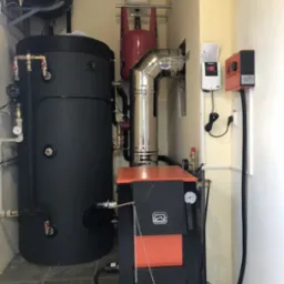 Kotłownia z czarnym zasobnikiem ciepłej wody, piecem na paliwo stałe z pomarańczowym frontem, instalacją hydrauliczną i sterownikami na ścianie.