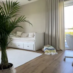 Jasne, beżowe zasłony z delikatnym marszczeniem, wiszące przy oknie w pokoju z drewnianą podłogą, kremową ścianą w łuski i białym łóżkiem z szufladami, z widoczną rośliną w wiklinowym koszu...