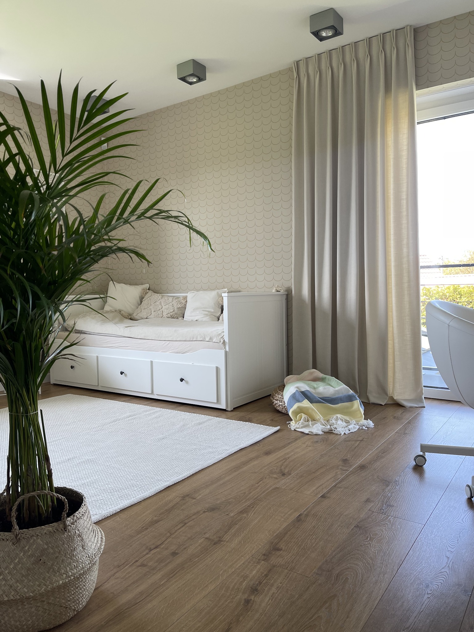 Jasne, beżowe zasłony z delikatnym marszczeniem, wiszące przy oknie w pokoju z drewnianą podłogą, kremową ścianą w łuski i białym łóżkiem z szufladami, z widoczną rośliną w wiklinowym koszu...