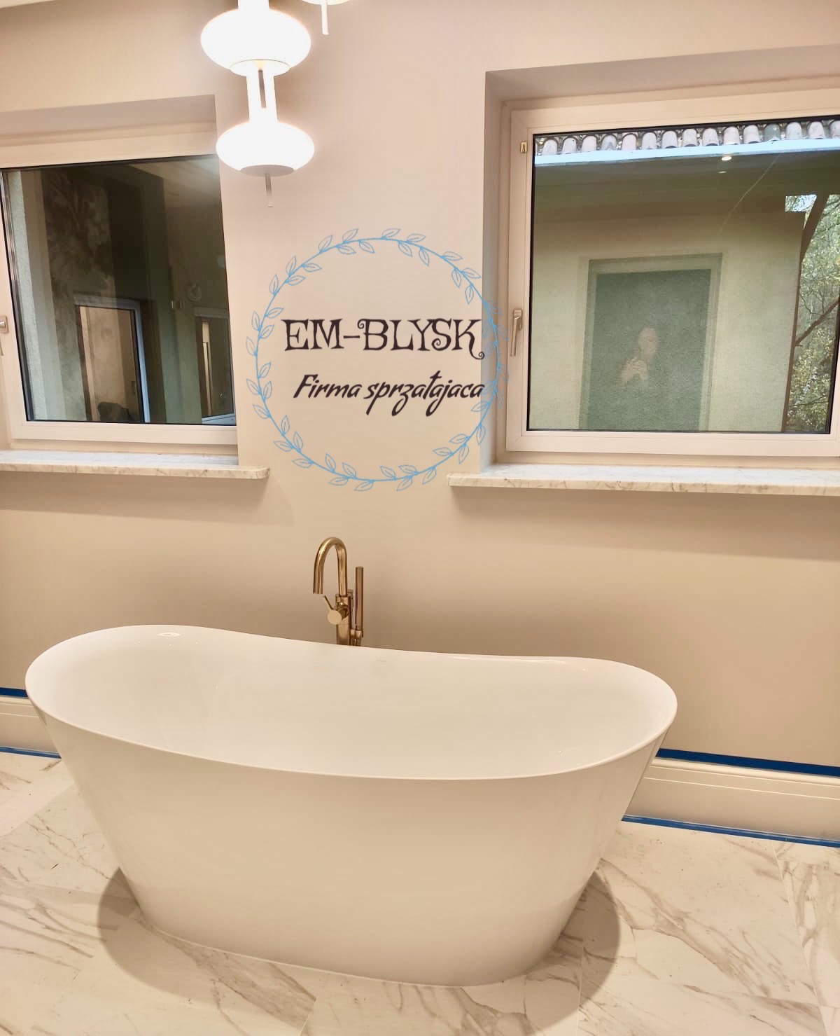 Nowoczesna, wolnostojąca biała wanna z złotą baterią w jasnej łazience z marmurową podłogą, logo firmy sprzątającej 'EM-BŁYSK' na ścianie.