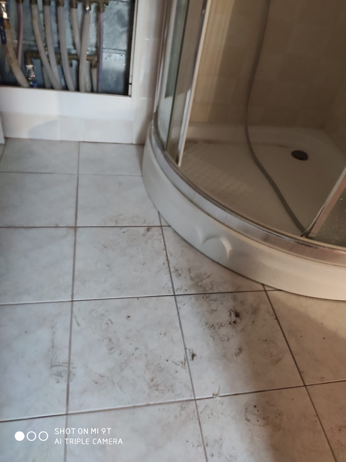 Brudna podłoga z płytek w łazience, widoczna kabina prysznicowa i rury doprowadzające wodę, wymagająca dokładnego wyszorowania.