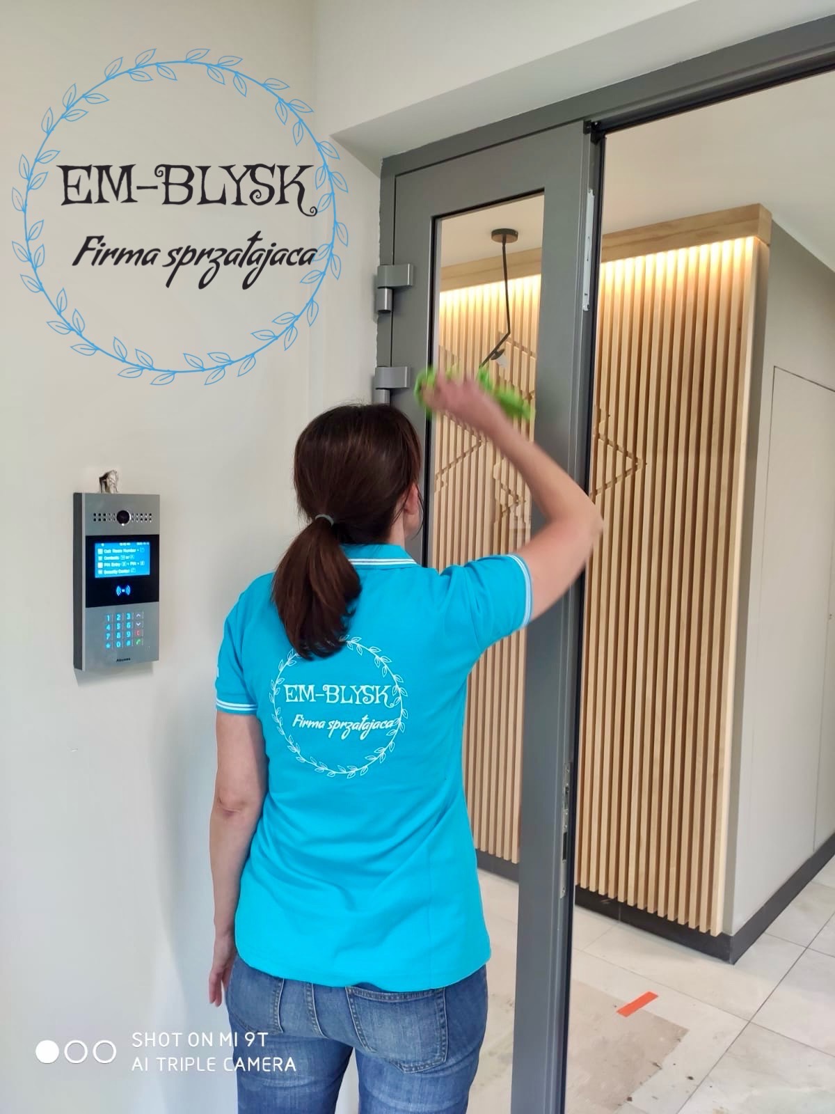 Kobieta w niebieskiej koszulce z logo firmy sprzątającej 'EM-BŁYSK' czyści szklane drzwi wejściowe do biura, w tle widoczny panel z numerami i drewniane panele dekoracyjne.