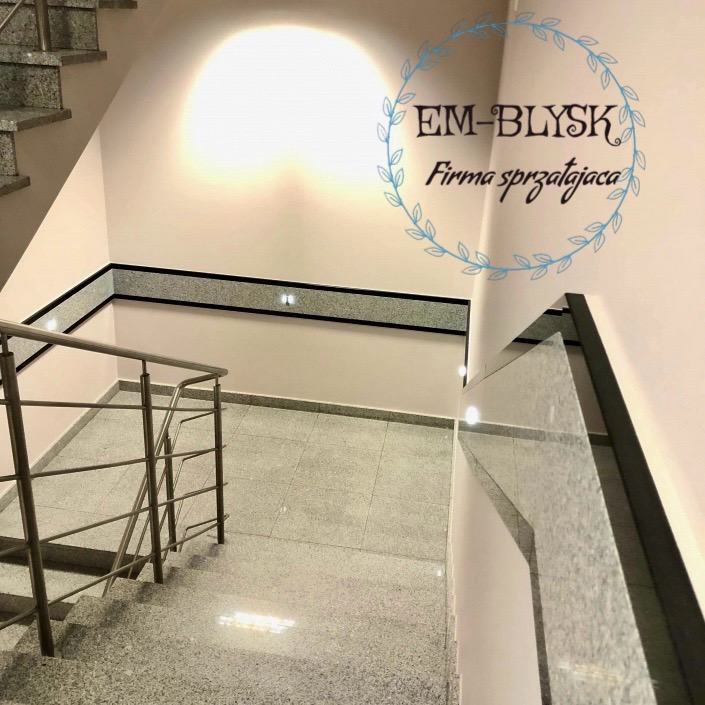 Klatka schodowa w budynku biurowym po sprzątaniu, widoczne błyszczące, szare płytki na podłodze i schodach, jasne ściany z ciemnym pasem dekoracyjnym i logo firmy sprzątającej na ścianie.