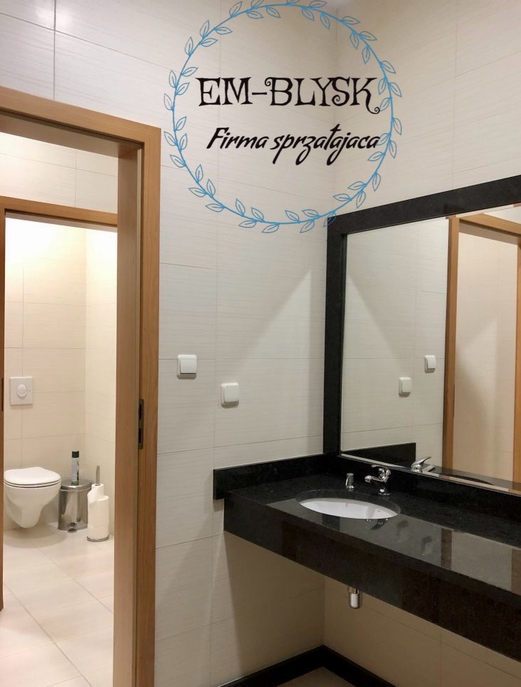 Czysta łazienka z toaletą i umywalką z czarnym blatem, widoczne logo firmy sprzątającej 'EM-BŁYSK' na ścianie.