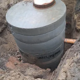 Betonowy zbiornik na szambo częściowo zakopany w ziemi, widoczna pokrywa i rura odpływowa, ziemia wokół wykopu jest błotnista.