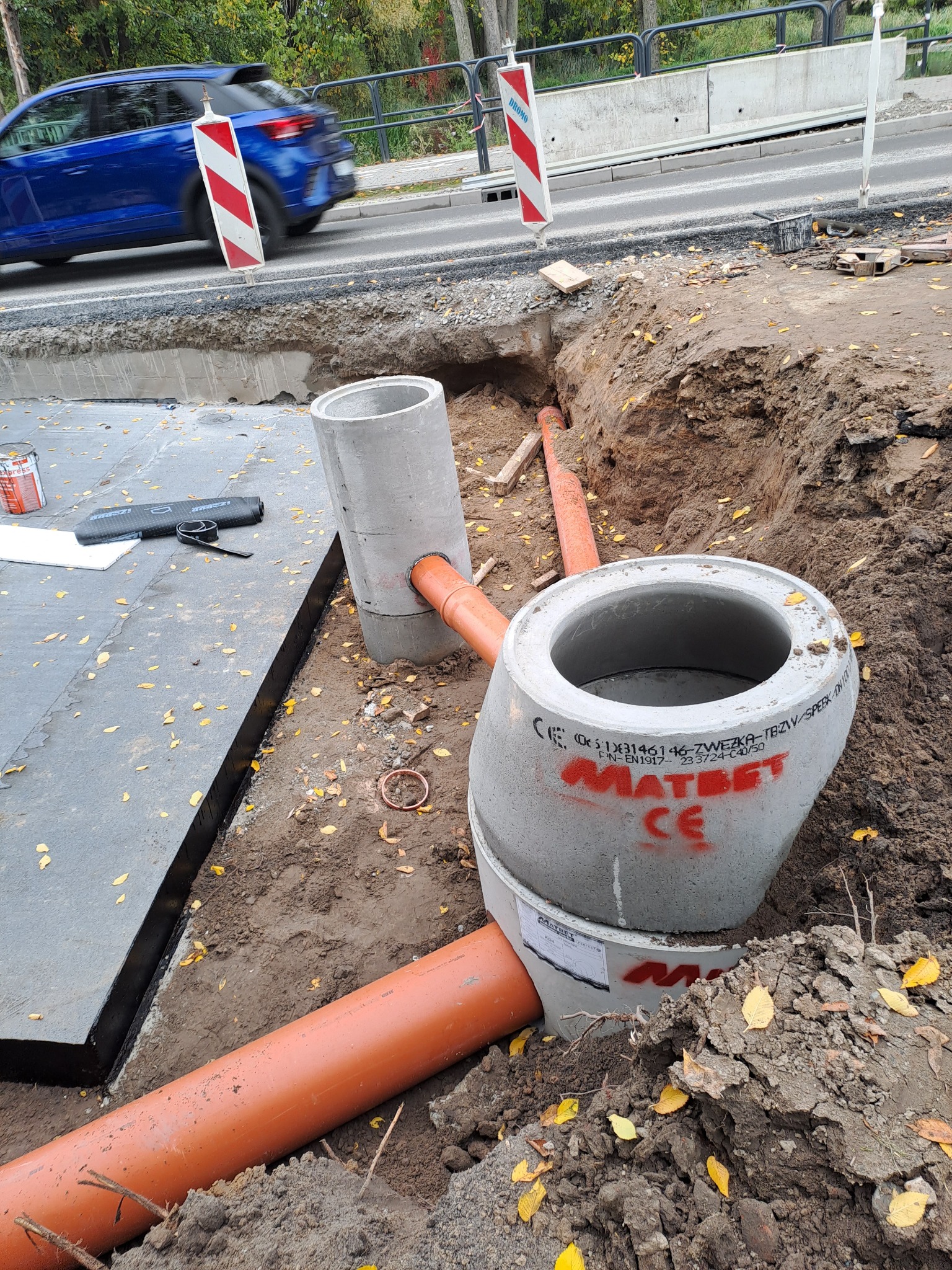 Wykonanie przyłącza kanalizacyjnego: betonowe studzienki z rurami PCV w wykopie, widoczne oznaczenia producenta Matbet, na pierwszym planie żółte liście, w tle ulica z barierami i przejeżdżający...