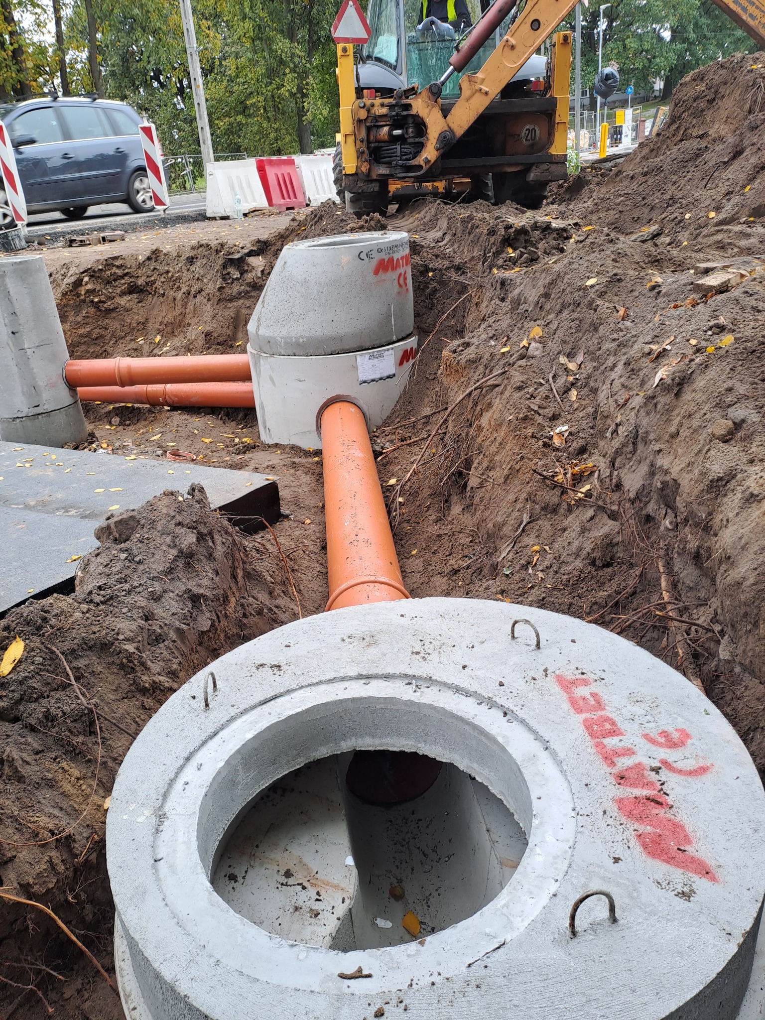 Montaż betonowych elementów kanalizacyjnych z pomarańczowymi rurami łączącymi, widoczny wykop i koparka w tle.