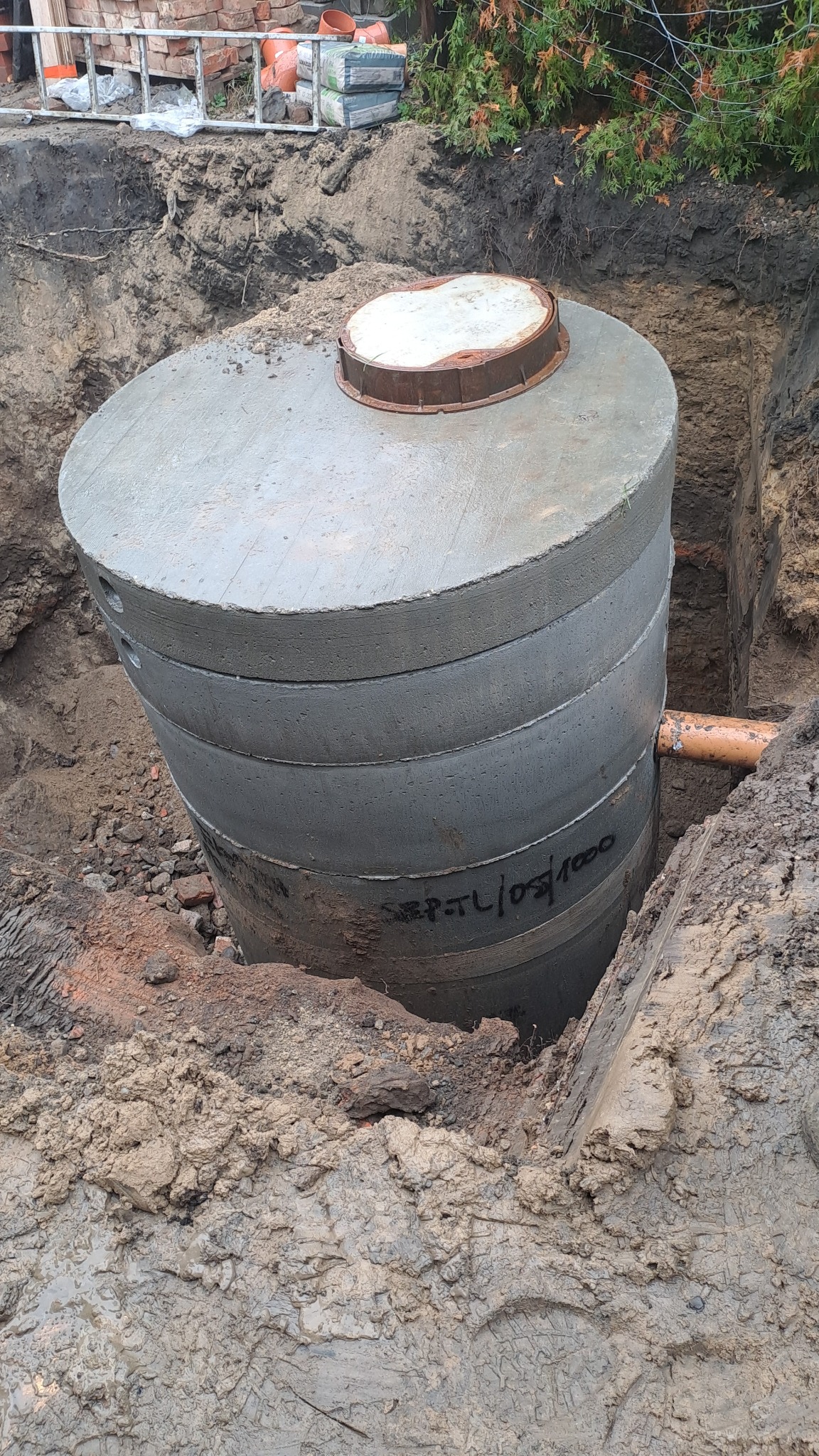 Betonowy zbiornik szamba z pokrywą, częściowo zagłębiony w ziemi, z widocznym podłączeniem rury kanalizacyjnej i napisem 'SEPT.TL/05/1000'