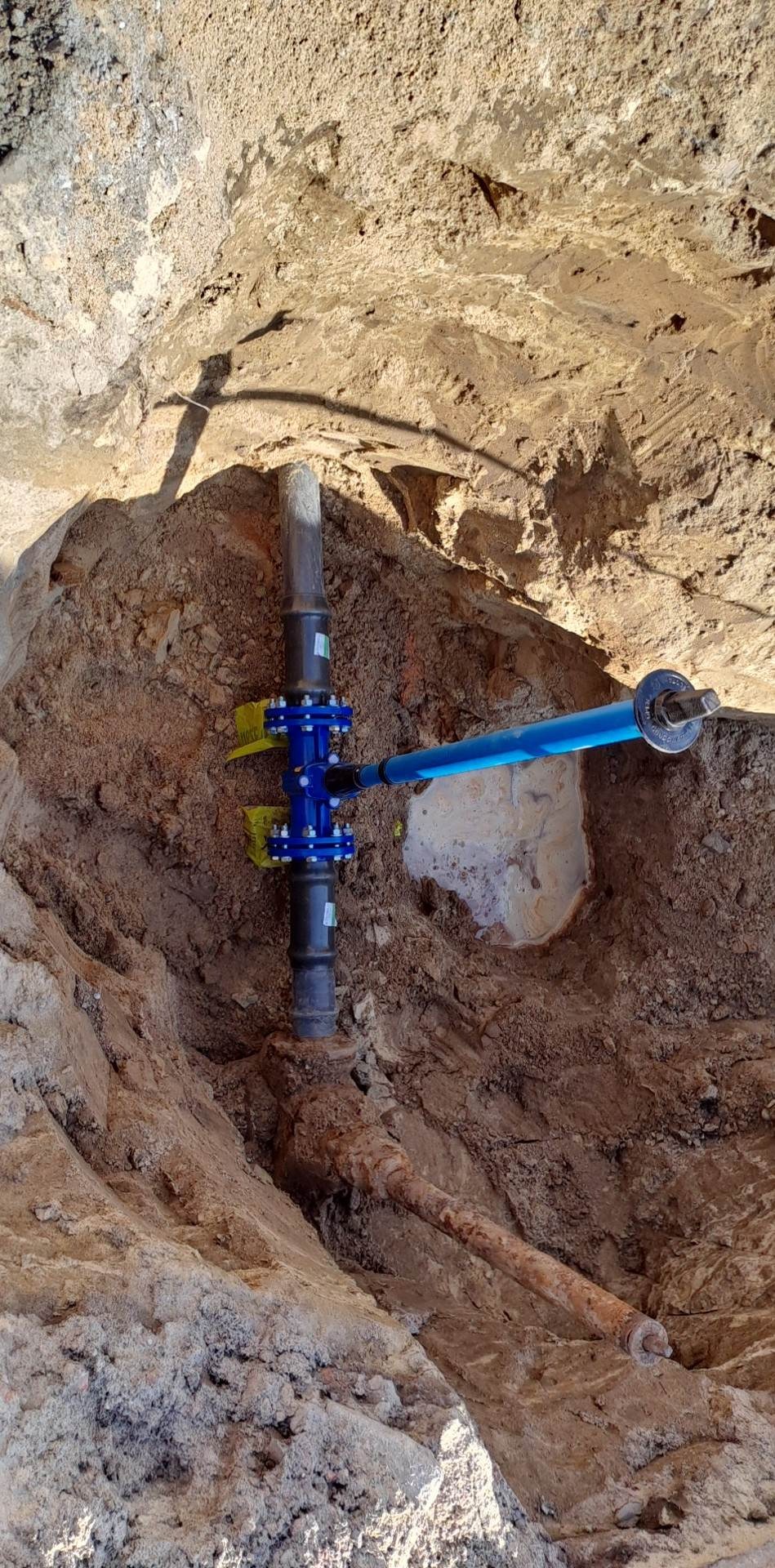 Wykonanie nowego przyłącza wodociągowego: pionowa rura z niebieskim zaworem i niebieską rurą odchodzącą w bok, w wykopie ziemnym z widoczną starą, rdzewiejącą rurą.