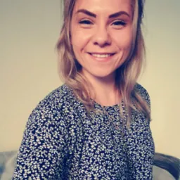 Portret uśmiechniętej blondynki w kwiecistej bluzce na tle kremowej ściany, lekko z boku.