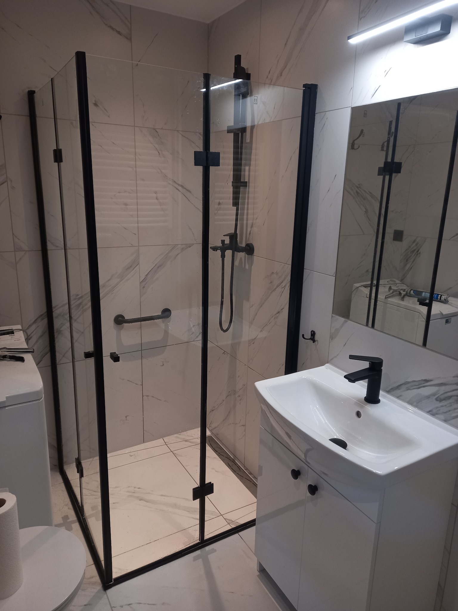 Nowoczesna kabina prysznicowa z czarnymi profilami i armaturą, biała umywalka z szafką w łazience wykończonej marmurowymi płytkami.