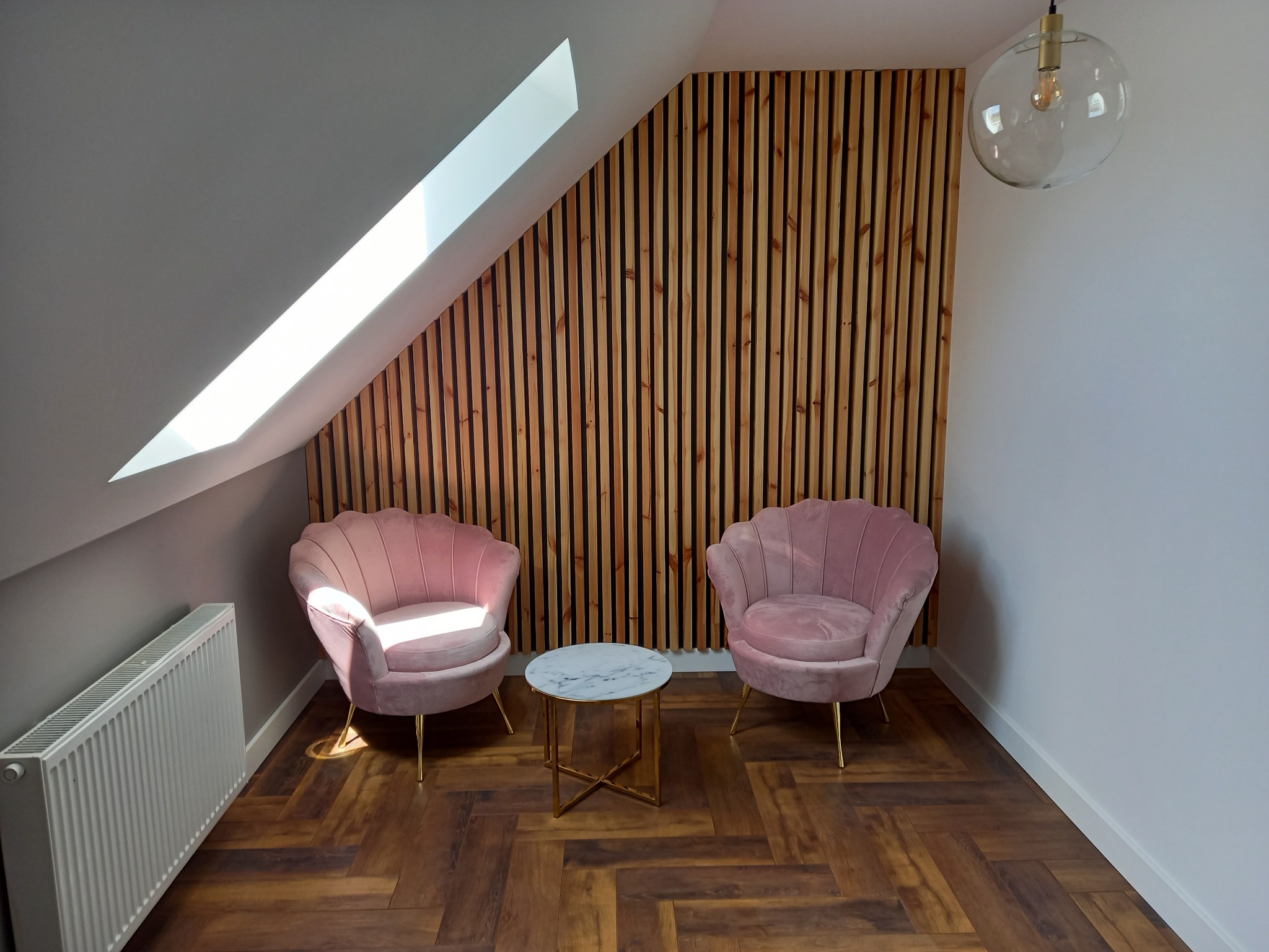 Poddasze z drewnianą boazerią, dwoma różowymi fotelami muszelkami, marmurowym stolikiem kawowym i lampą wiszącą, oświetlone naturalnym światłem z okna dachowego.