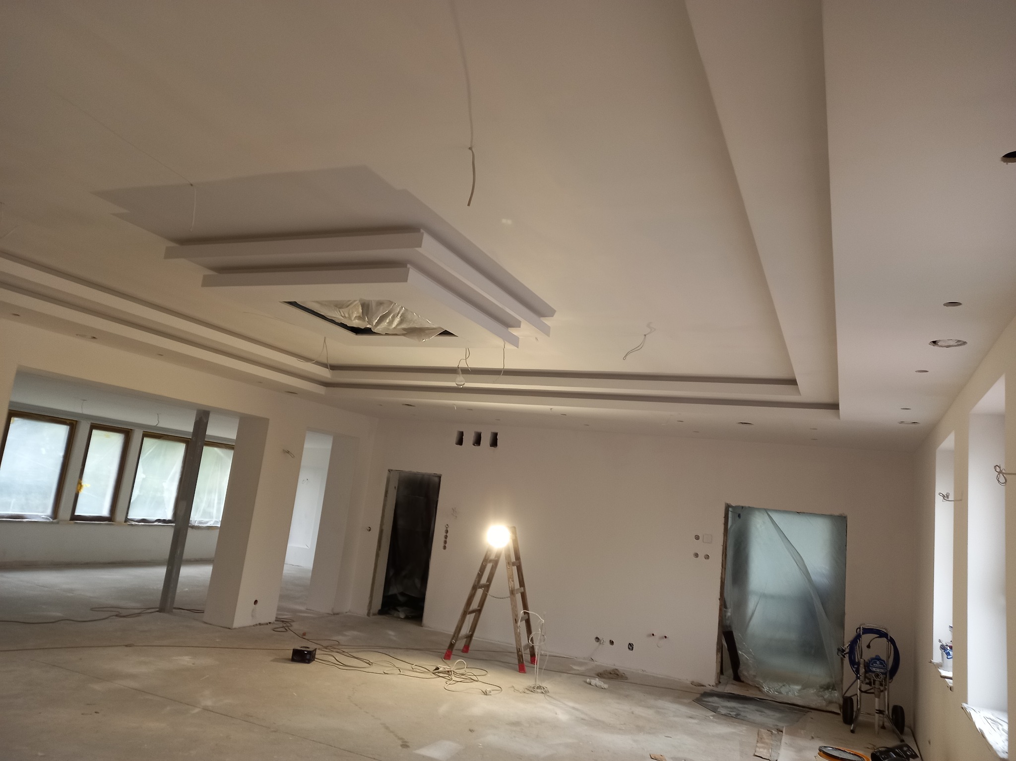 Wnętrze w trakcie wykończenia: białe ściany, dekoracyjny sufit z podwieszanymi elementami, drabina z halogenem, zakryte folią drzwi i okna, agregat malarski oraz widoczne instalacje elektryczne.