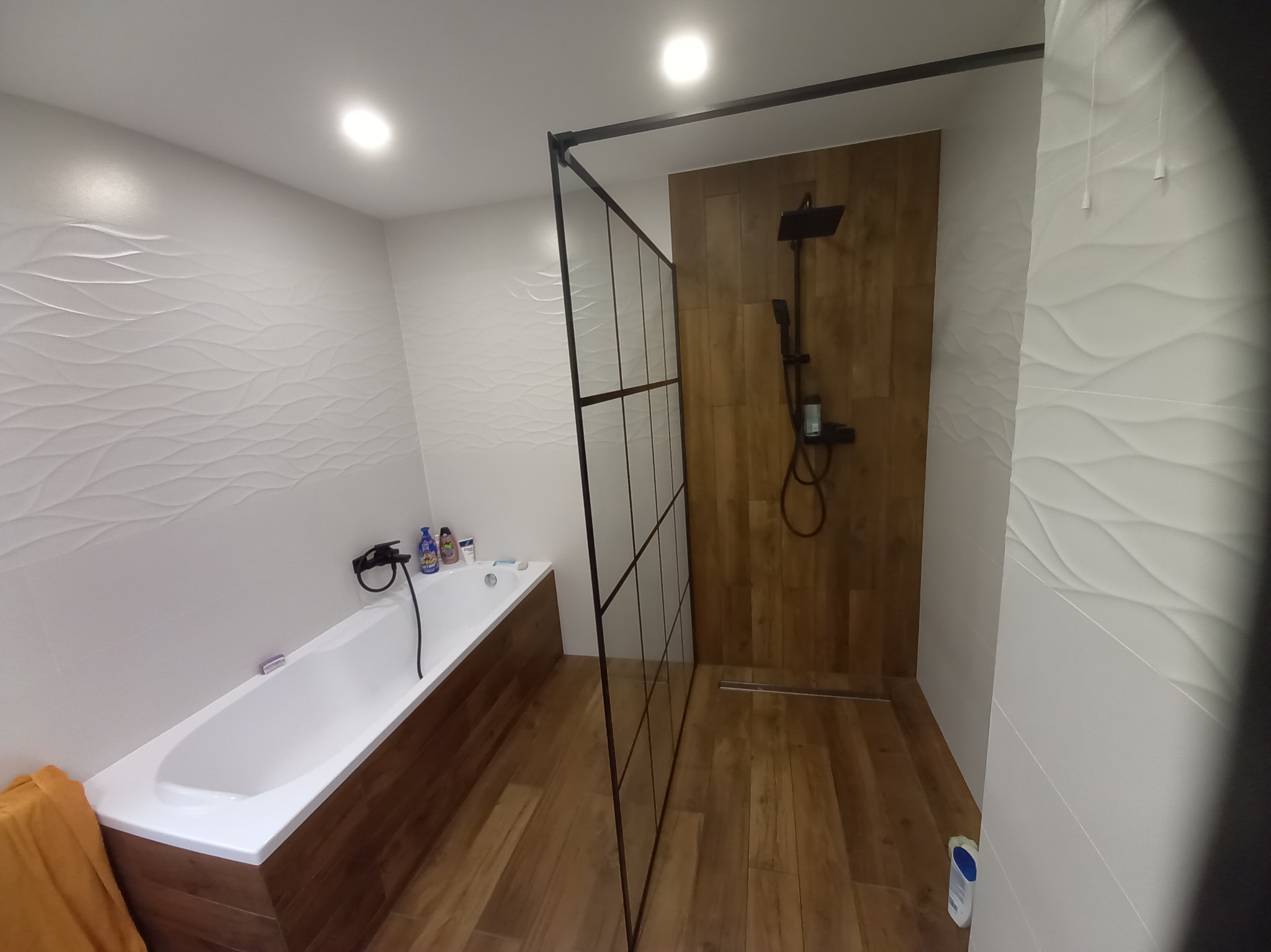 Nowoczesna łazienka w Rzędowie z wanną obudowaną drewnem i kabiną prysznicową typu walk-in z czarnymi profilami, wykończona płytkami imitującymi drewno i białymi płytkami z falistą strukturą.