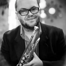 Portret czarno-biały muzyka w okularach, trzymającego trąbkę, z uśmiechem w tle dekoracyjne elementy wnętrza.