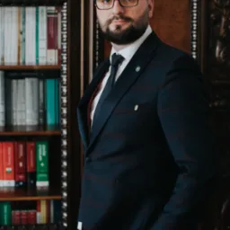 Portret mężczyzny w garniturze na tle drewnianego regału z książkami, z zegarkiem na nadgarstku i przypinkami w klapie.