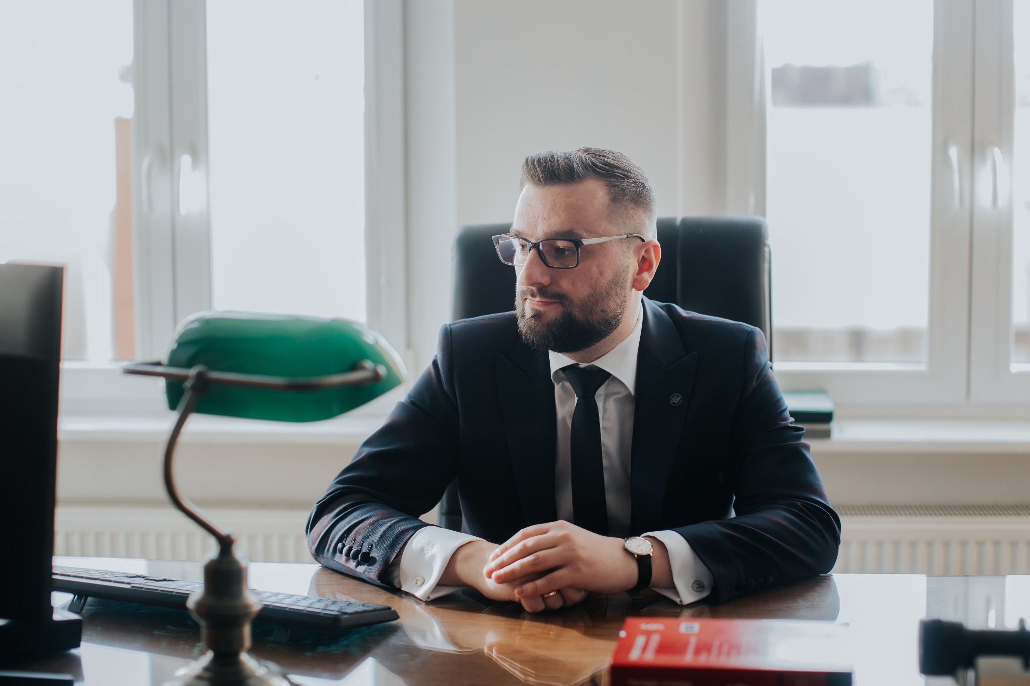 Portret mężczyzny w garniturze siedzącego za biurkiem z zieloną lampką bankierską, spoglądającego w bok, z rękami złożonymi na blacie.
