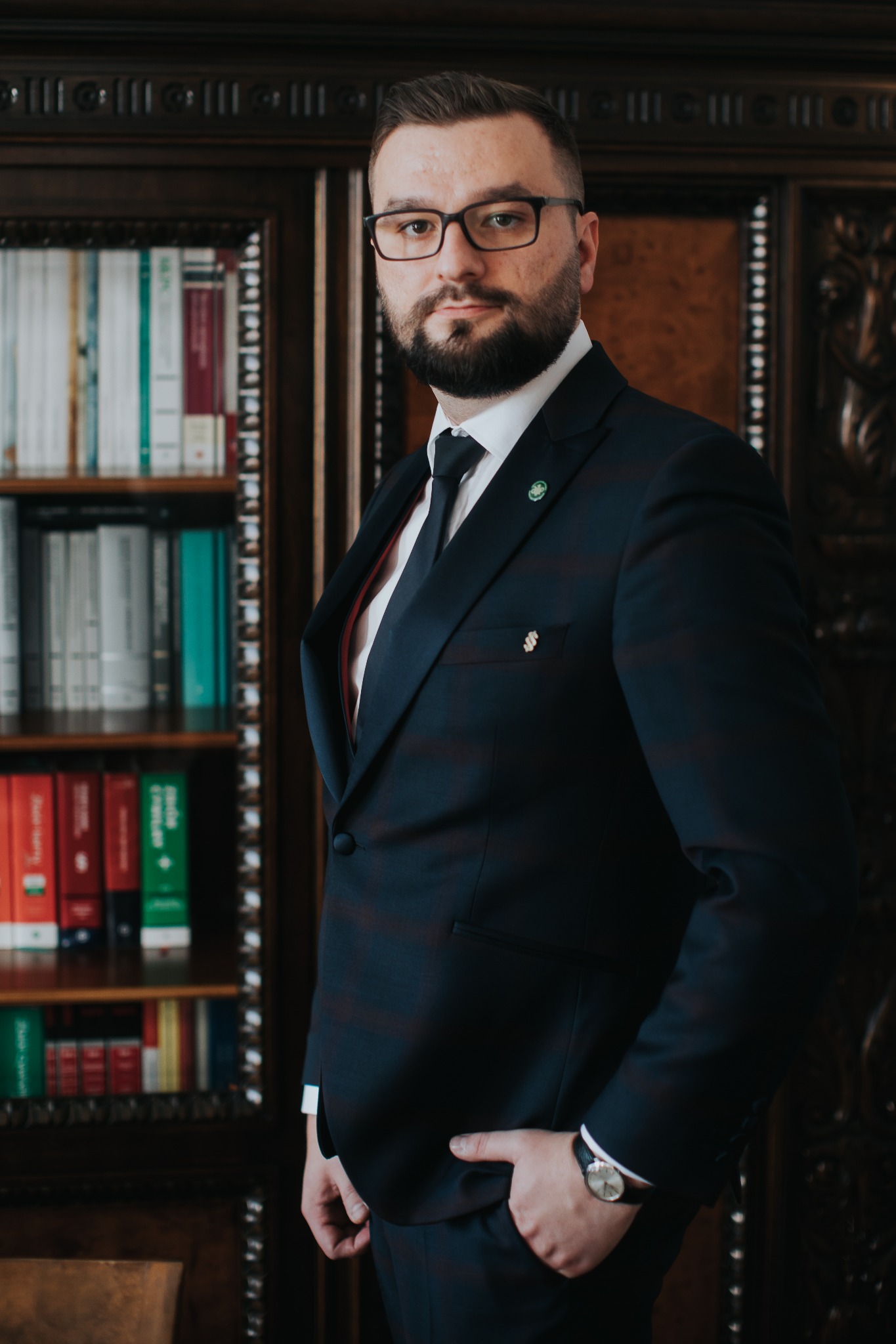 Portret mężczyzny w garniturze na tle drewnianego regału z książkami, z zegarkiem na nadgarstku i przypinkami w klapie.