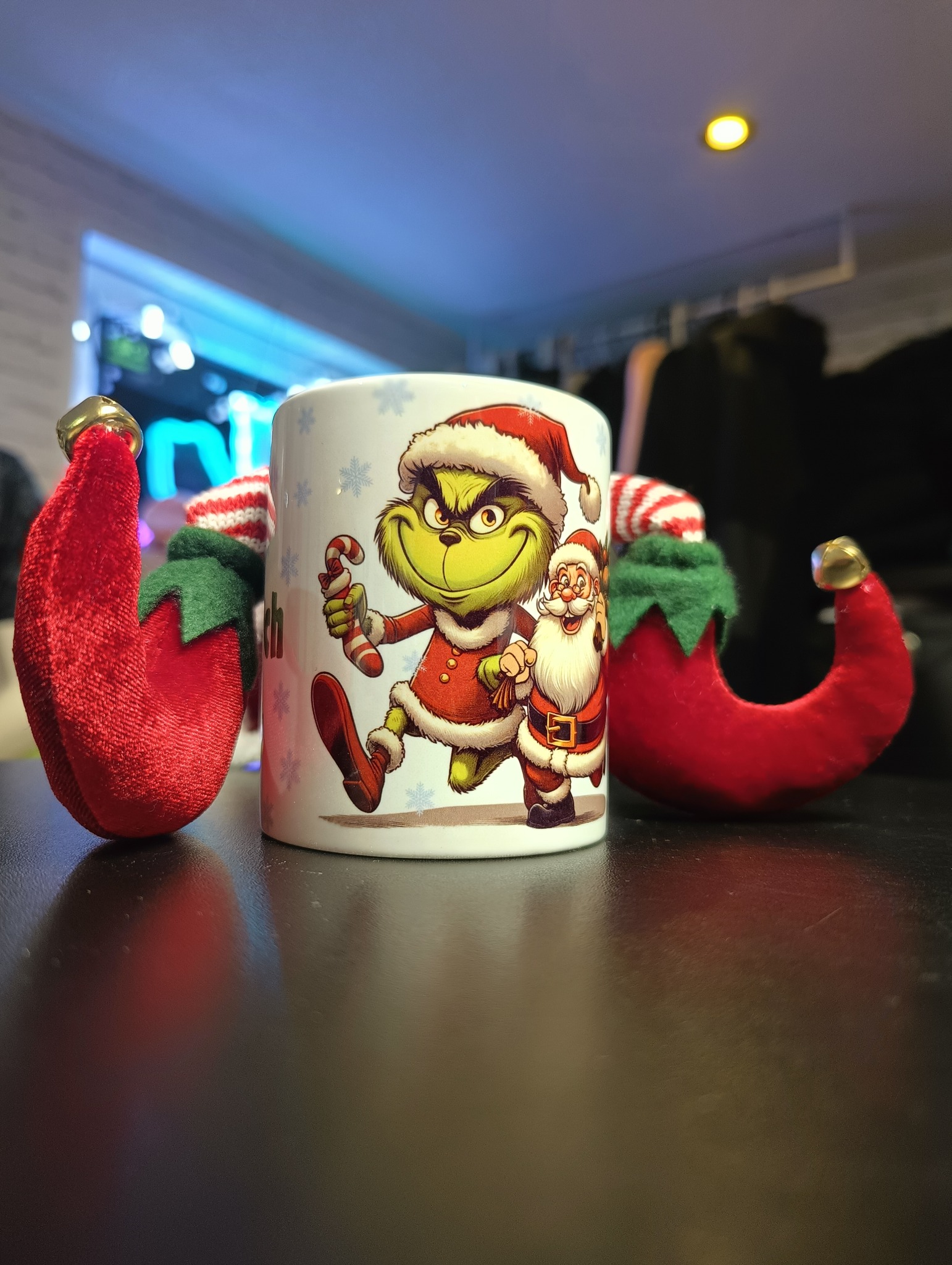 Ceramiczny kubek z nadrukiem Grincha i Świętego Mikołaja w świątecznych strojach, otoczony czerwonymi elfimi czapeczkami z dzwoneczkami, na ciemnym błyszczącym blacie.