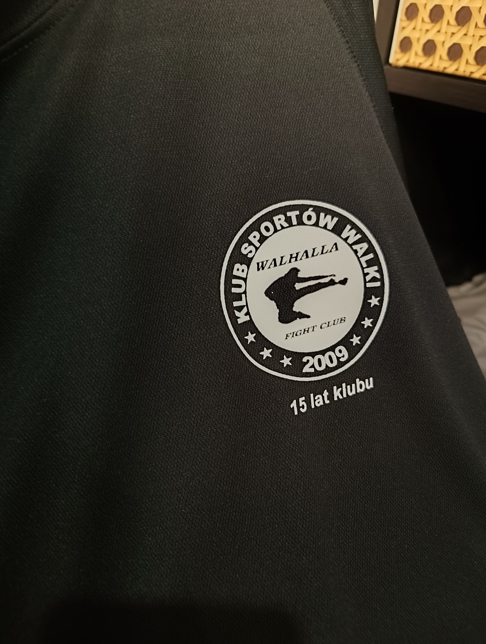 Czarna koszulka sportowa z okrągłym, białym nadrukiem logo klubu sportów walki 'Walhalla Fight Club' z sylwetką zawodnika i rokiem założenia 2009, świętującego 15-lecie.