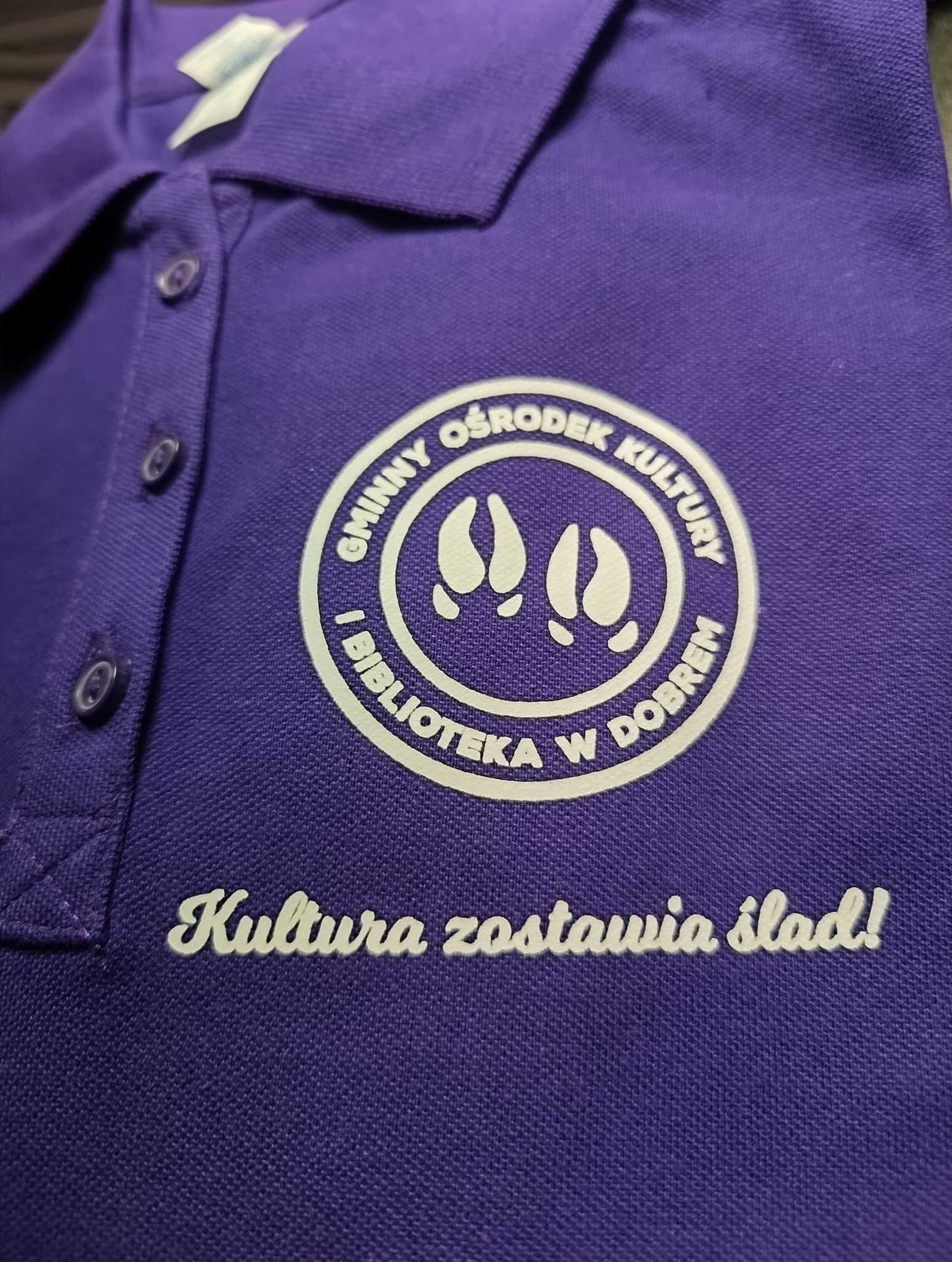 Fioletowa koszulka polo z białym nadrukiem logo Gminnego Ośrodka Kultury i Biblioteki w Dobrem oraz hasłem 'Kultura zostawia ślad!'