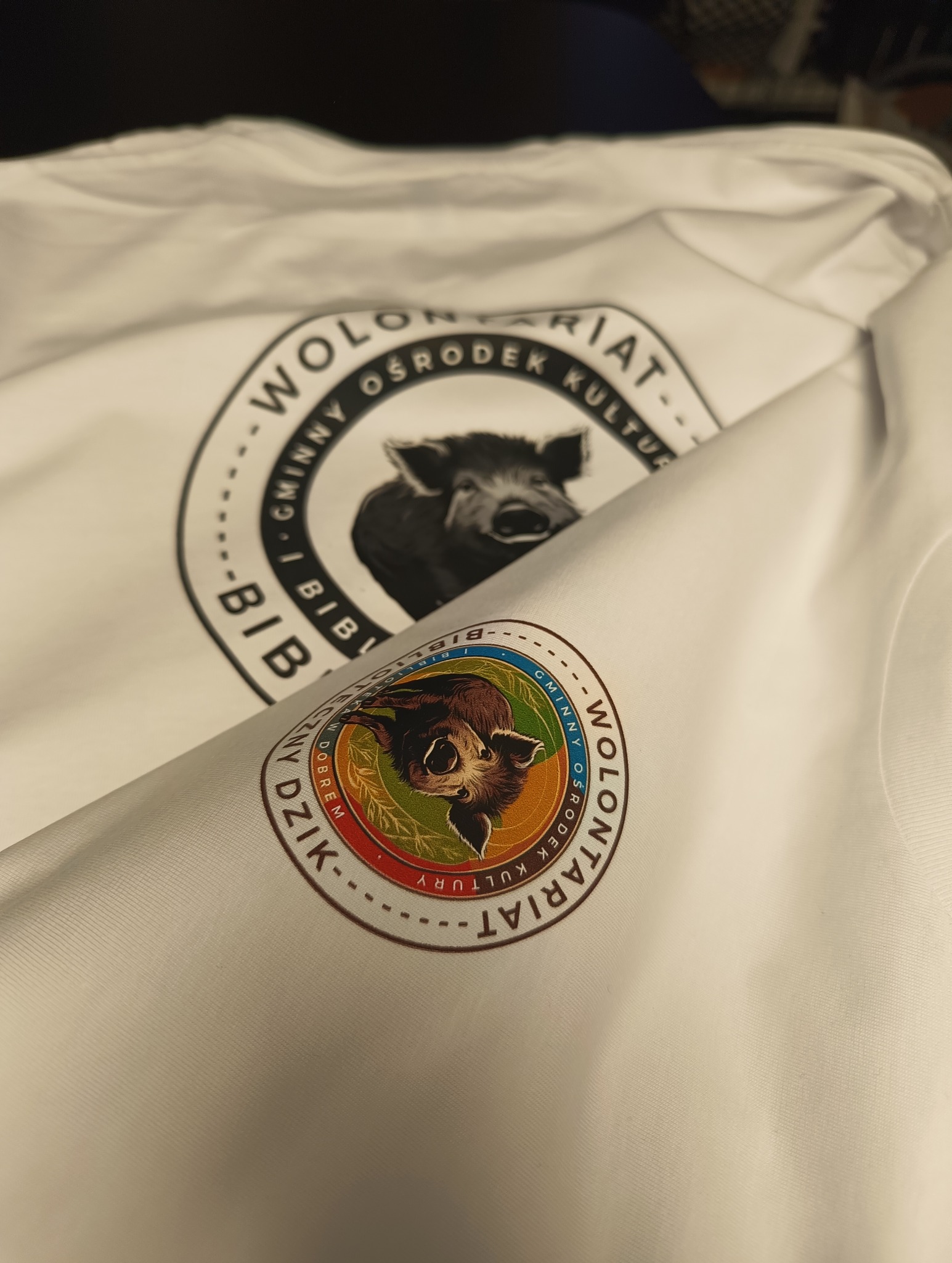 Biała koszulka z nadrukowanym logo przedstawiającym wizerunek dzika i nazwą 'Wolontariat Gminny Ośrodek Kultury w Dobrem' na piersi i na rękawie.